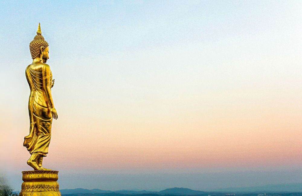 Golden standing Buddha statue at Khao Phra Tamnak Viewpoint in Pattaya, facing a scenic sunset horizon with panoramic views of the city and coastline.