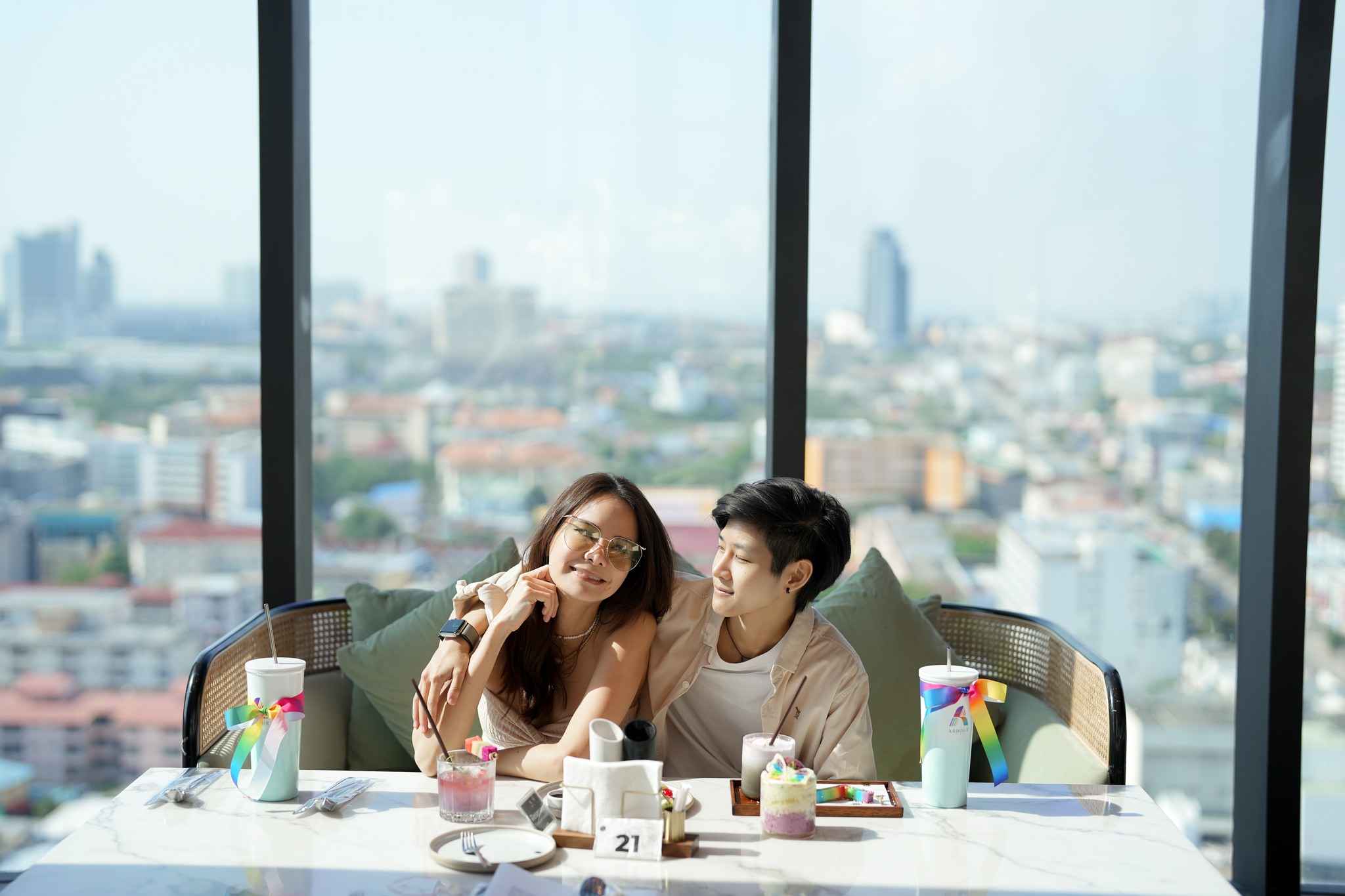 A couple enjoying a delightful meal at a rooftop dining area with a stunning view of the cityscape during the day. The table features colorful desserts and drinks with Pride-themed decorations.