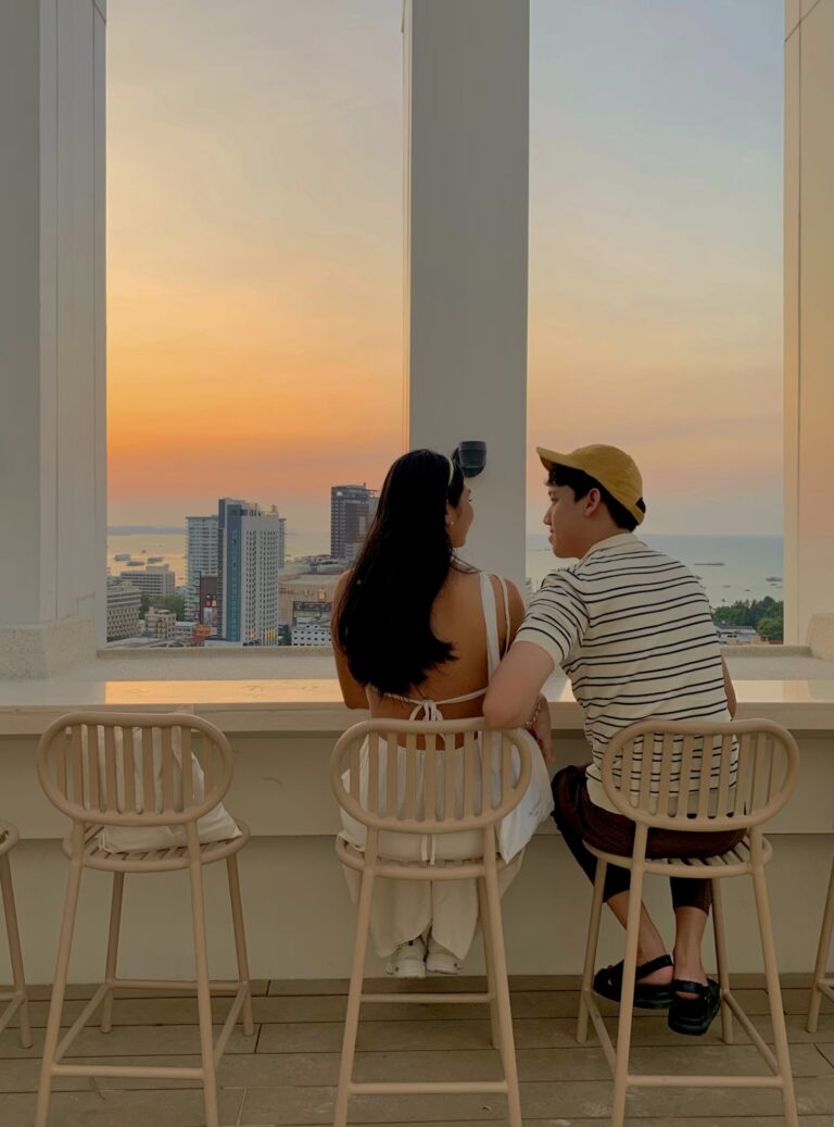 A couple sitting on barstools, enjoying a romantic sunset view from a rooftop in Pattaya, with the city and ocean in the background.