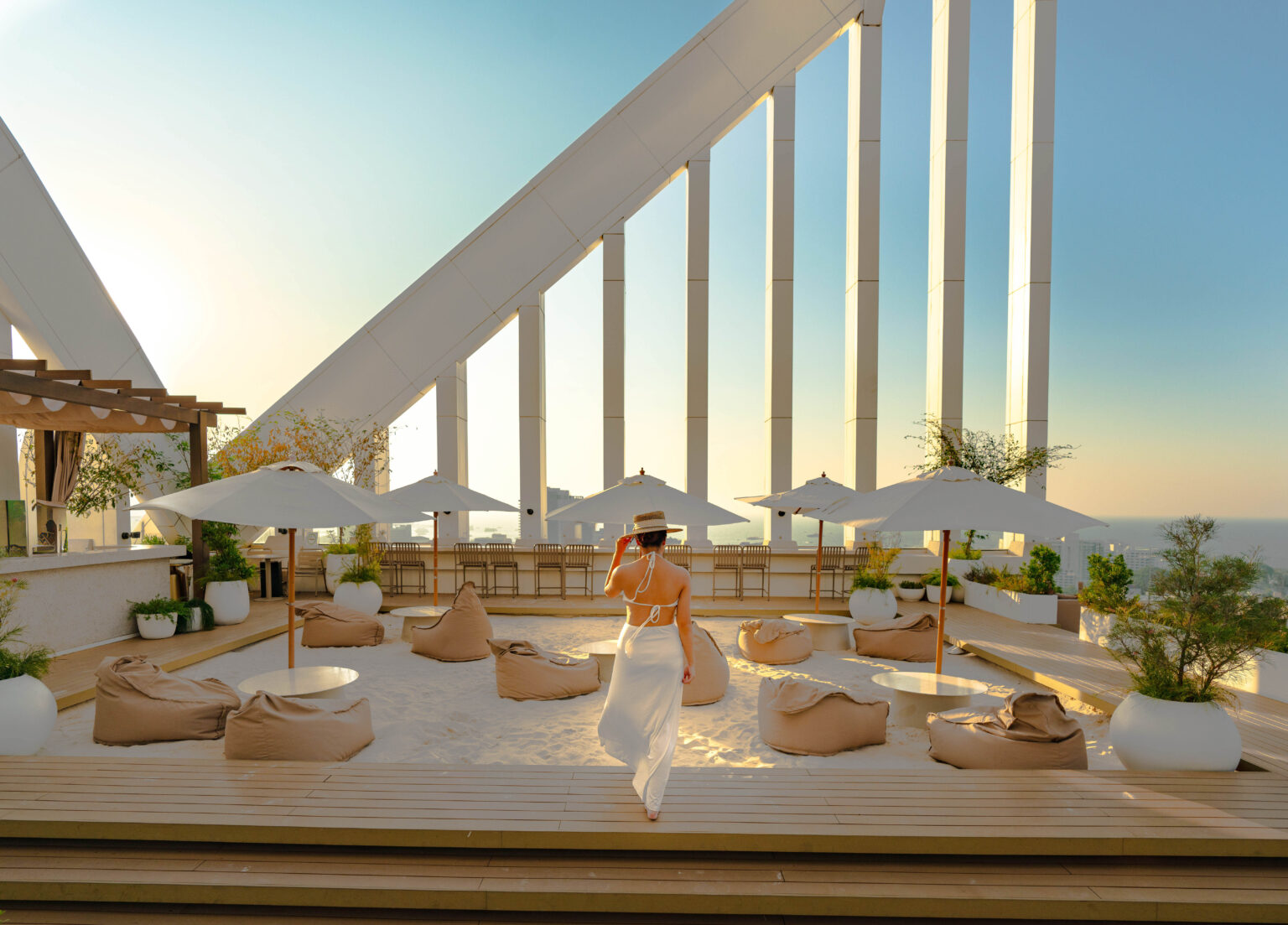 A woman in a white dress and straw hat walking toward a rooftop area with sand, bean bags, and umbrellas at sunset, featuring modern architecture and a scenic ocean view in Pattaya.