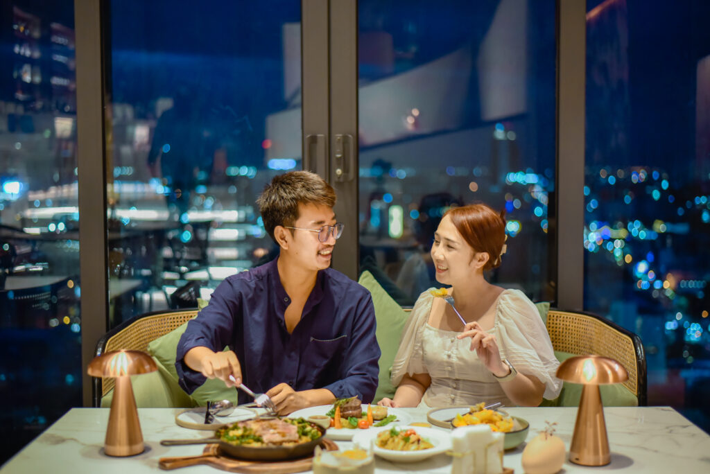 A couple enjoying a romantic dinner with gourmet dishes at a table by the window, overlooking a stunning city night view.