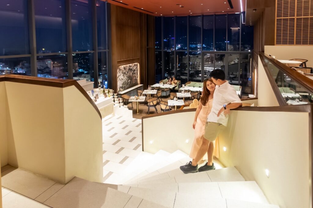 A couple enjoying a romantic moment on an elegantly lit staircase inside a sophisticated restaurant with panoramic night views of Pattaya city.