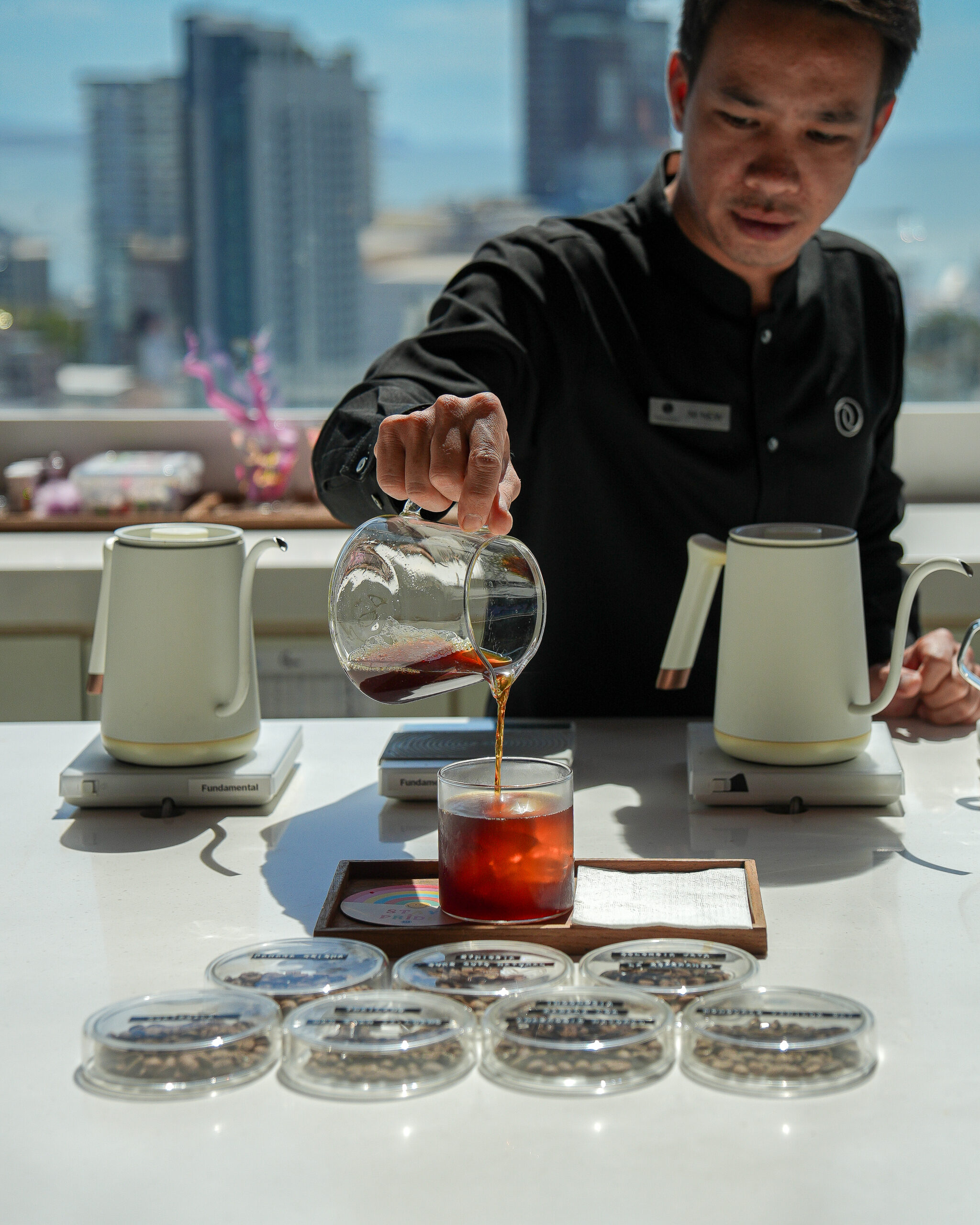 A barista pouring freshly brewed coffee into a glass at a modern cafe with a scenic city view in the background.