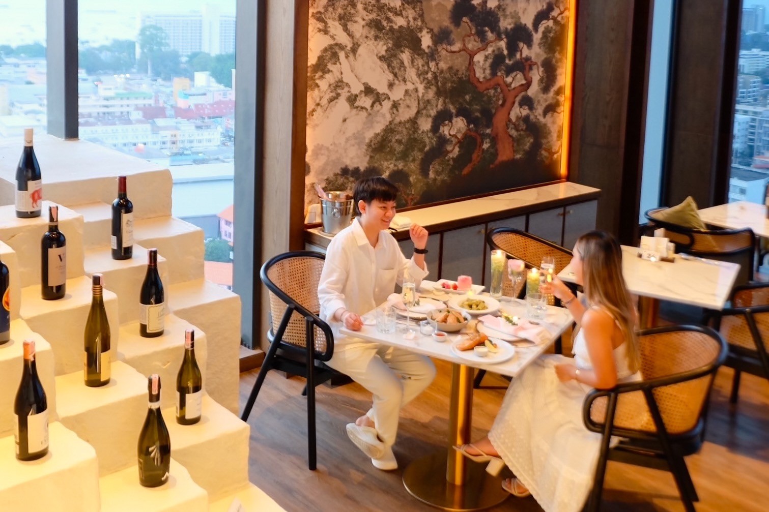 A couple enjoying an elegant dining experience at a table set with gourmet dishes and wine bottles, overlooking a vibrant cityscape through large windows.