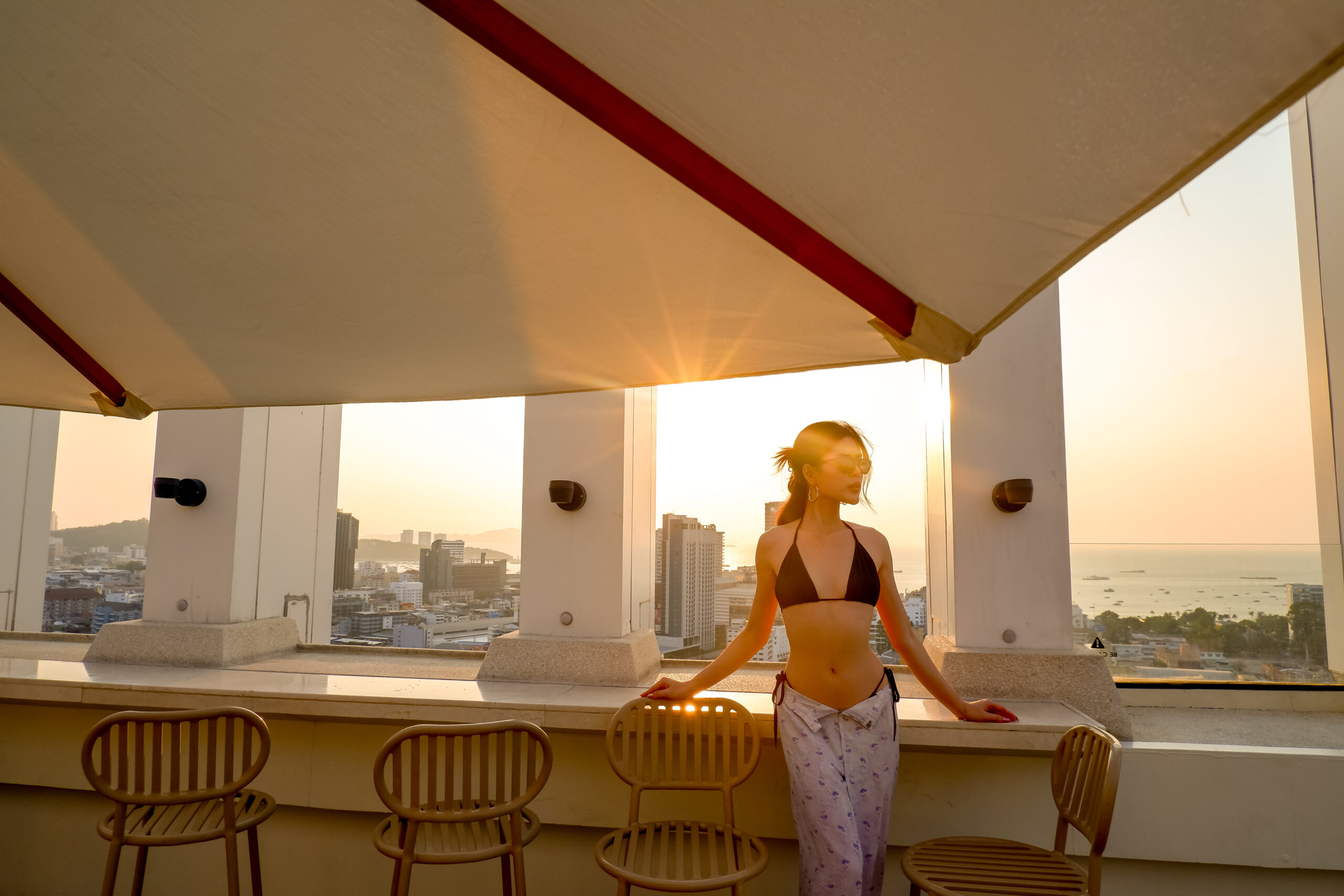 A woman in a black bikini top and patterned sarong standing at a rooftop bar during sunset, with a scenic view of Pattaya city and the ocean in the background.