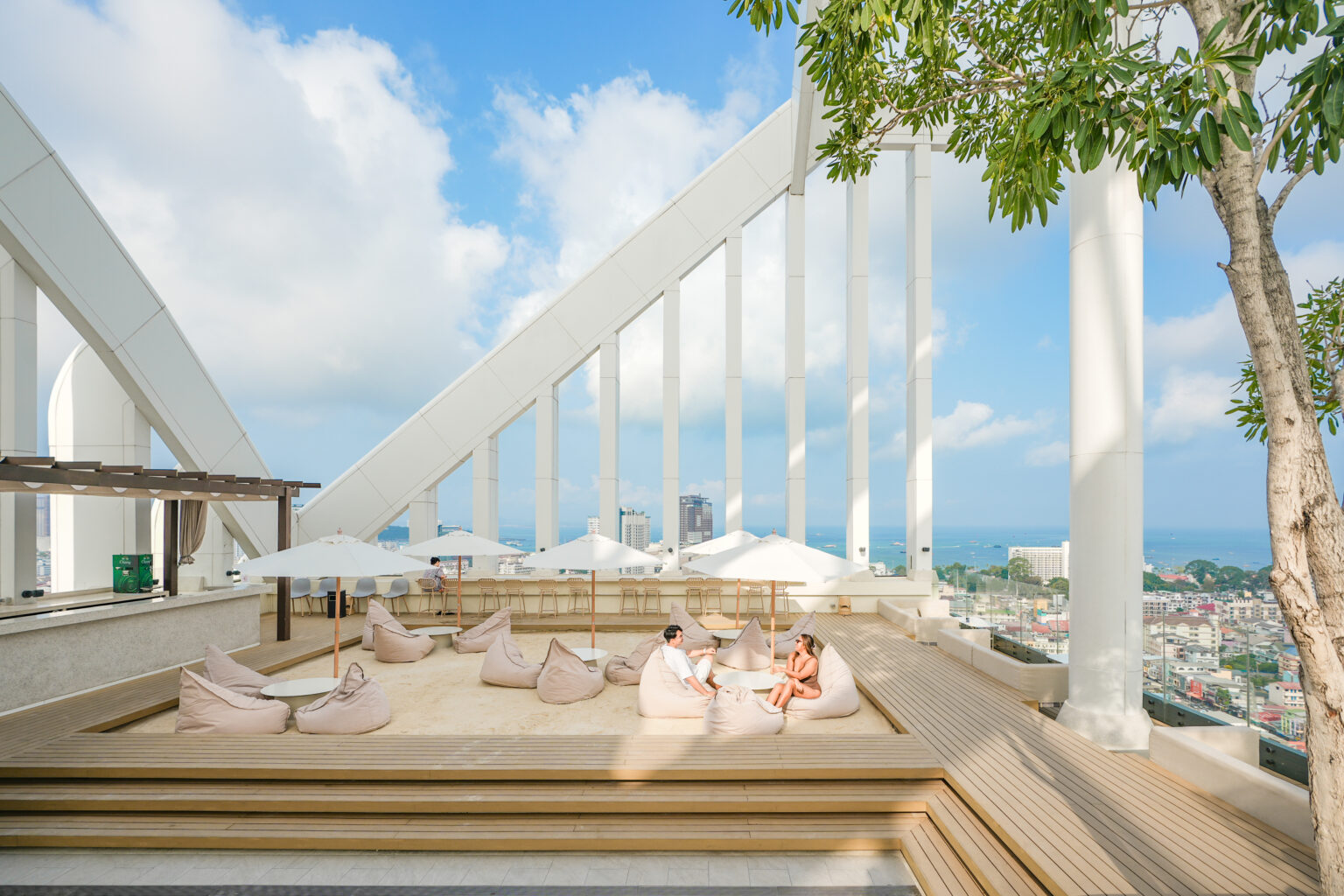 A couple relaxing on beanbags at a rooftop lounge with a modern architectural design, overlooking Pattaya city and the ocean under a sunny sky.