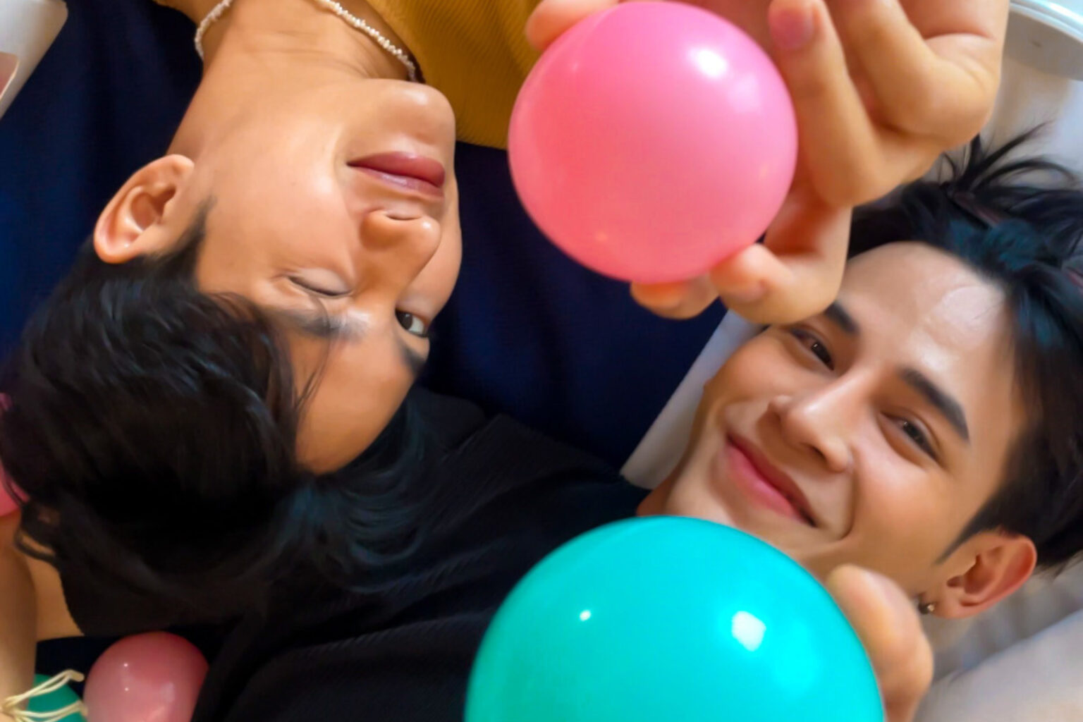 Two individuals lying on their backs and smiling while holding colorful pink and green balloons. The playful scene conveys warmth, joy, and connection.