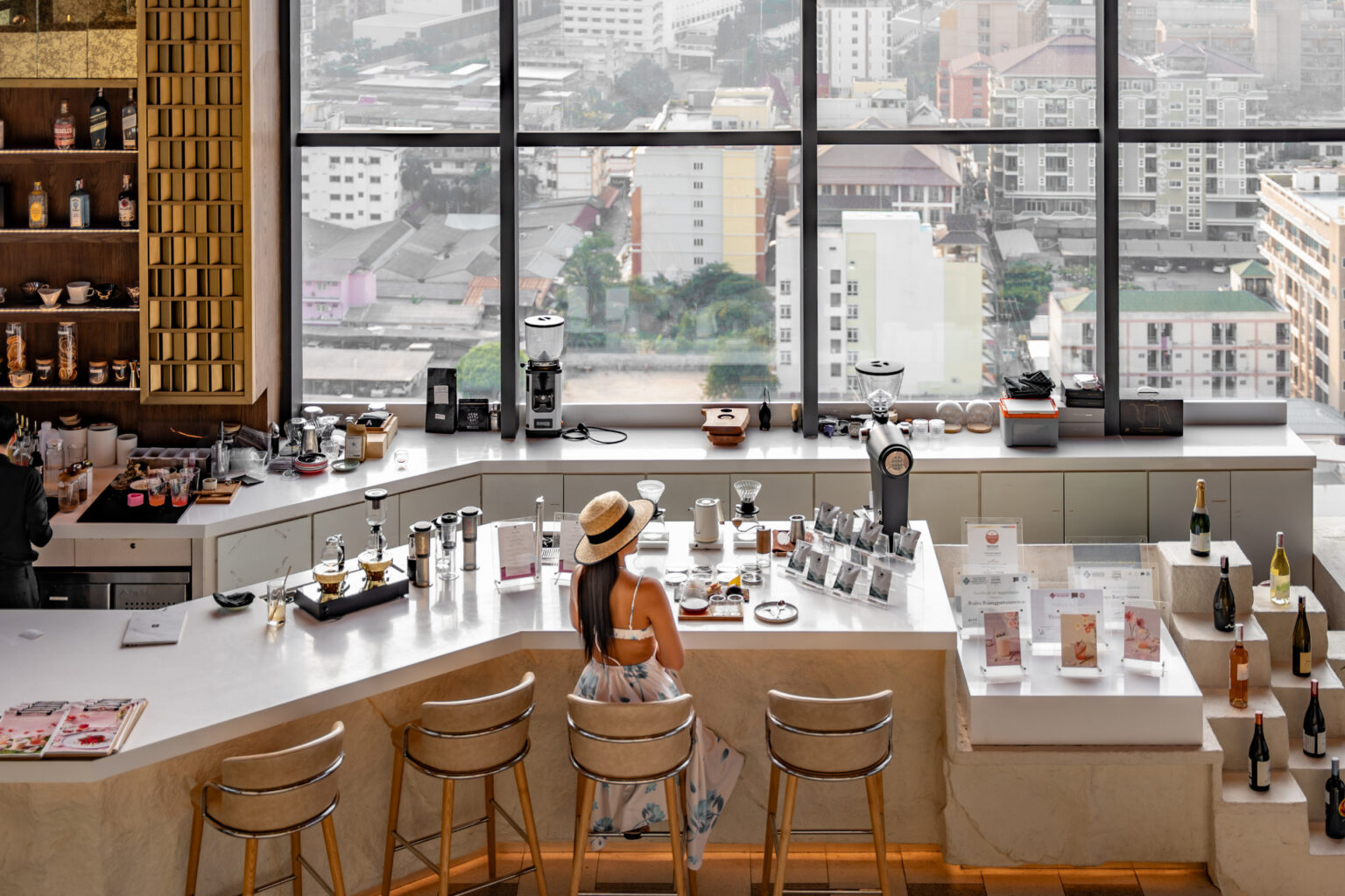 A stylish cafe with a panoramic city view, featuring a modern coffee bar, brewing equipment, and a woman enjoying her drink at the counter.