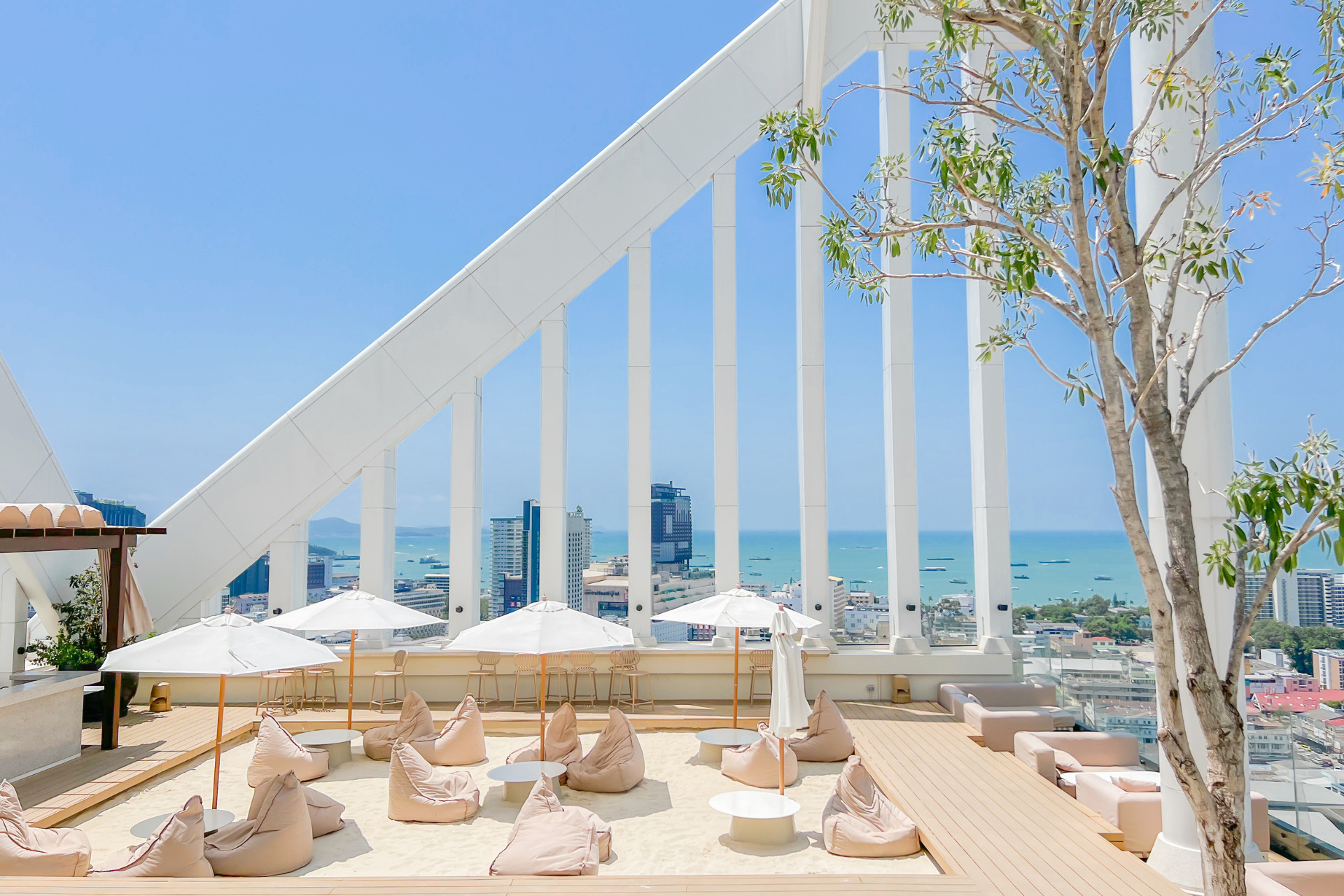 Bright and inviting rooftop bar with bean bag seating, white umbrellas, and panoramic views of Pattaya city and the ocean under a clear blue sky.