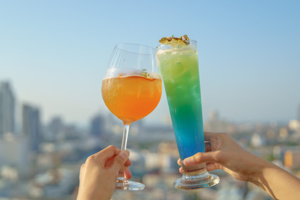 Close-up of two colorful cocktails, one orange and one blue-green, with a cityscape in the background under a clear sky.