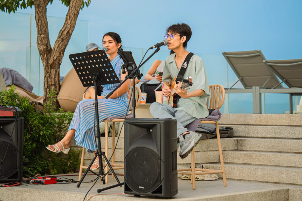 Acoustic duo performing live music on the rooftop, featuring a guitarist and singer with a clear blue sky in the background.