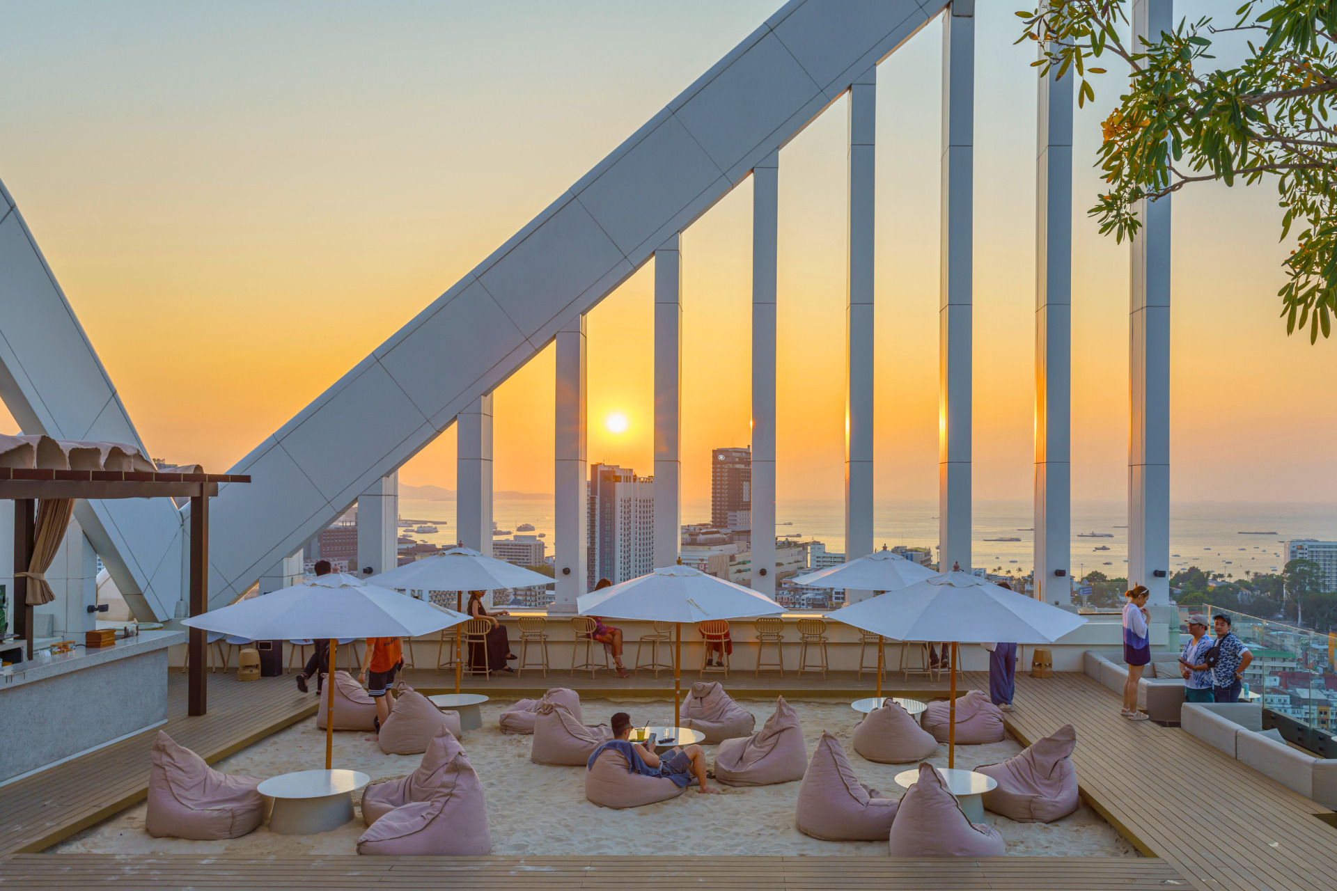 Elegant rooftop bar at sunset with bean bag seating, white umbrellas, and a panoramic view of Pattaya city and ocean.