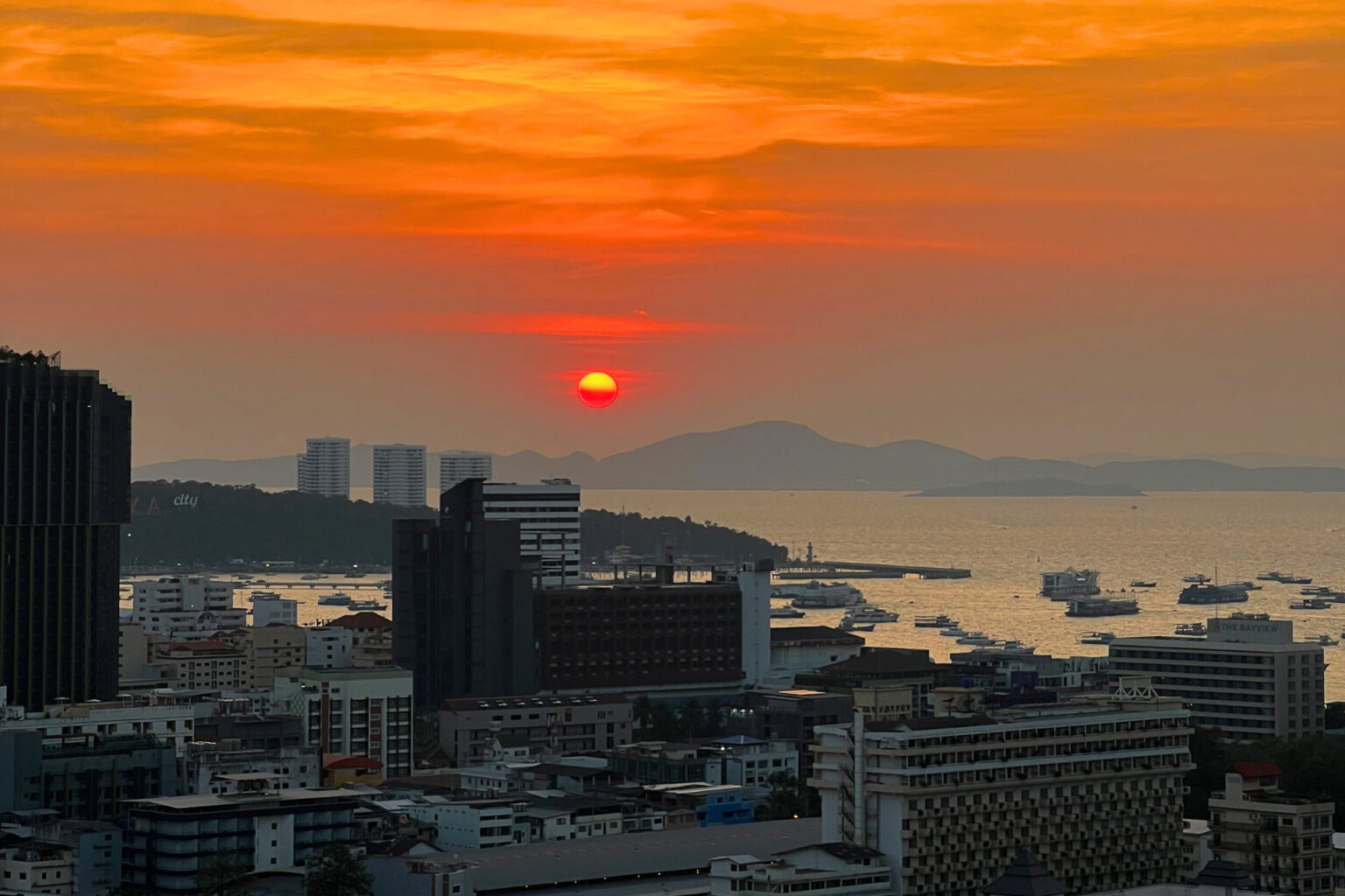 Vivid orange and red sunset over Pattaya city skyline with the ocean and distant hills in the background.