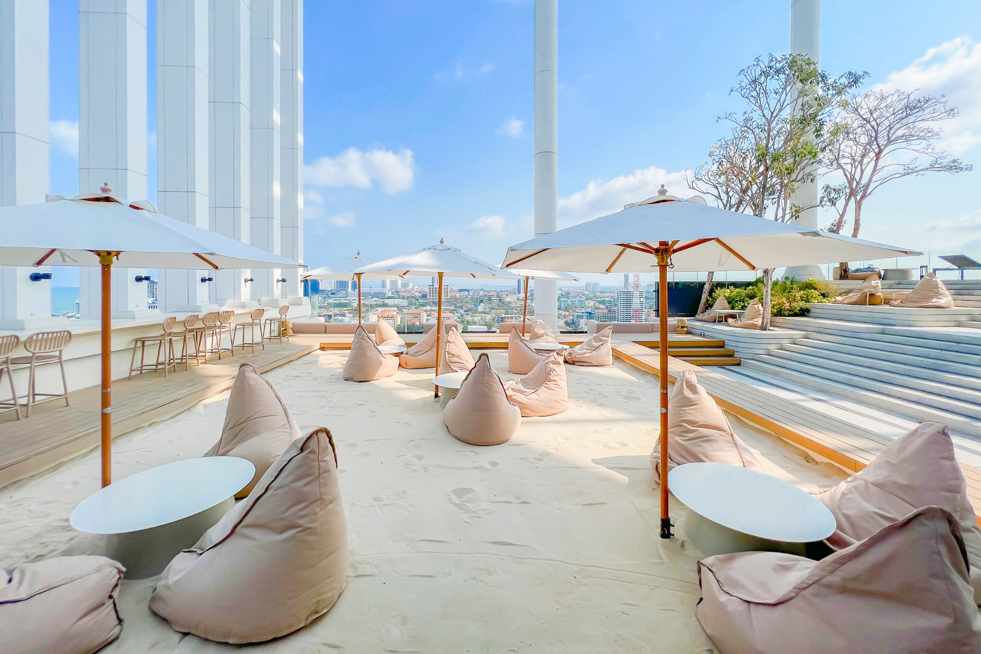 Rooftop bar at Arbour Hotel and Residence Pattaya with sandy seating area, bean bags, and white umbrellas overlooking Pattaya cityscape on a sunny day.