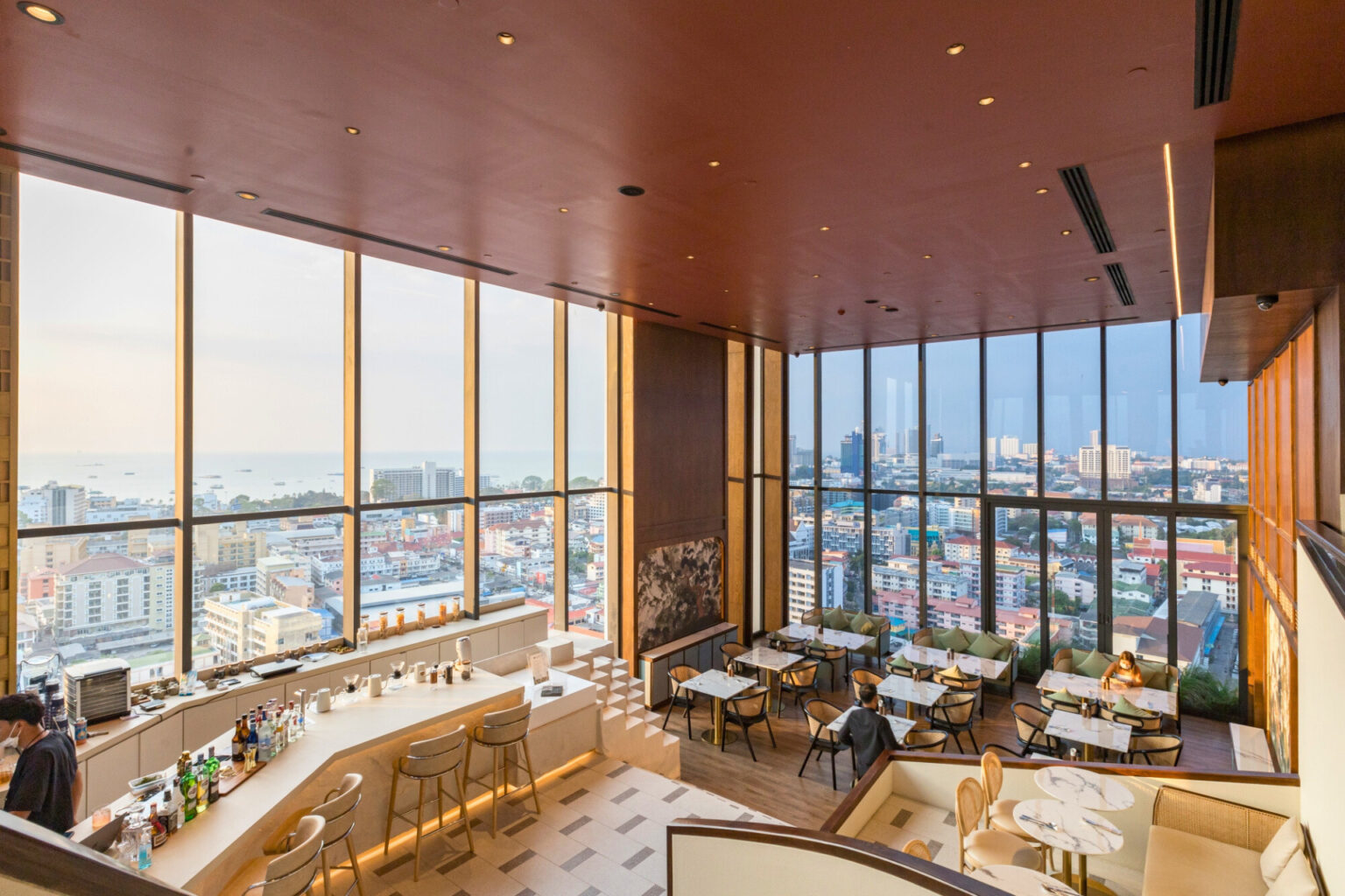 A modern cafe with floor-to-ceiling windows offering panoramic views of Pattaya city and the ocean, featuring chic seating and a stylish bar.