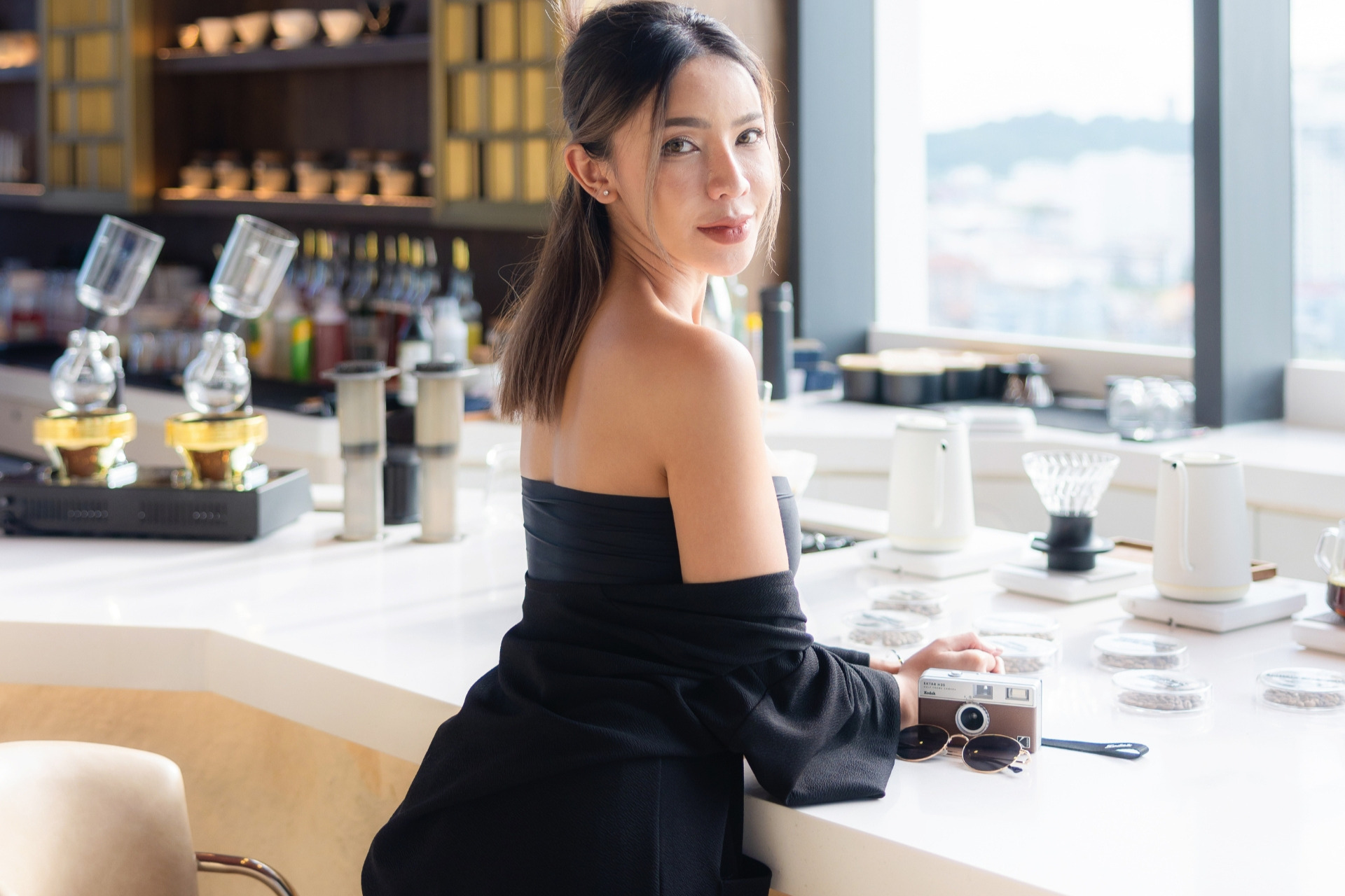 A fashionable woman in a black outfit seated at a coffee bar, surrounded by brewing equipment, with a relaxed yet stylish pose.