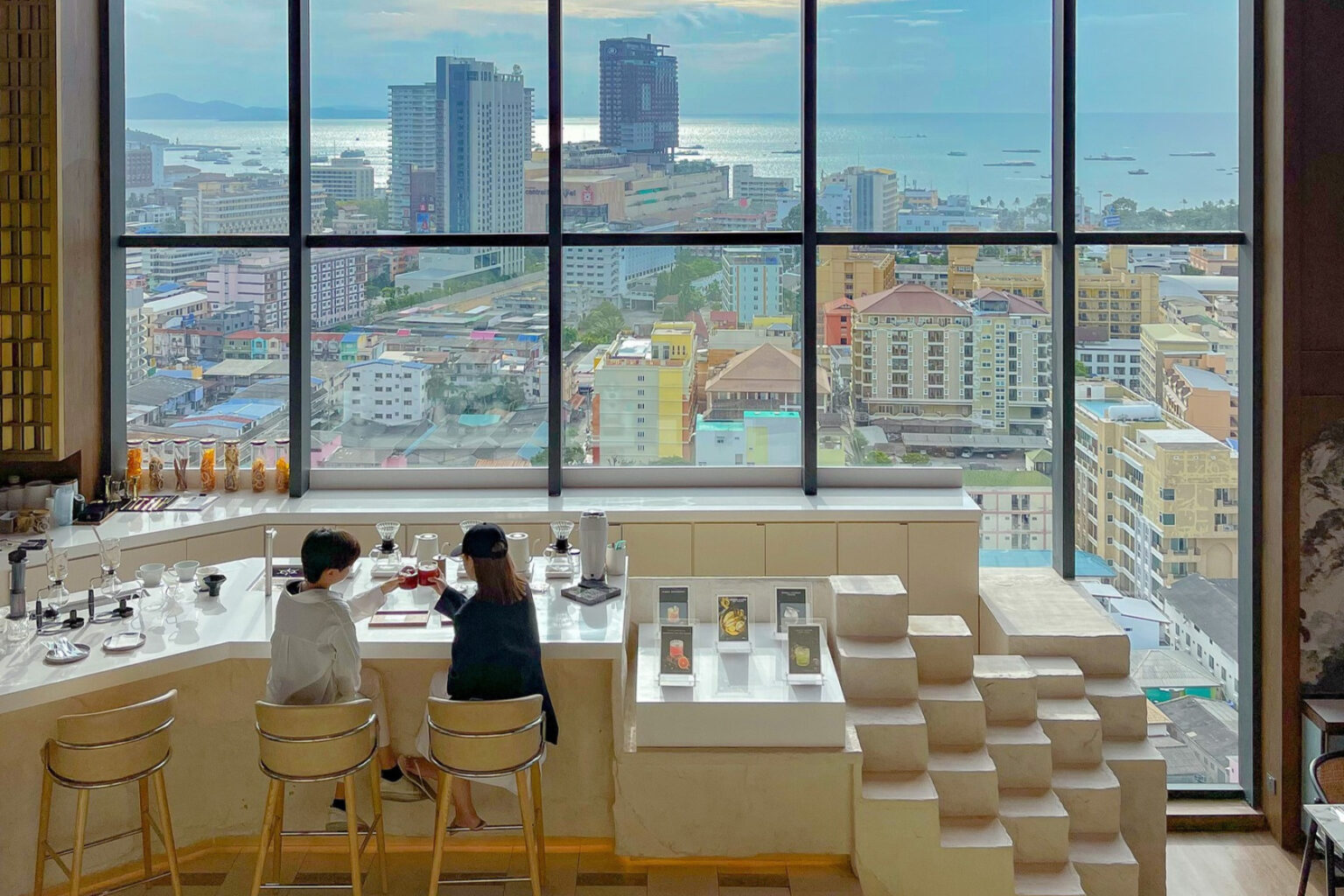 Two people enjoying coffee at a sleek bar with a panoramic view of Pattaya’s skyline and ocean through large floor-to-ceiling windows.