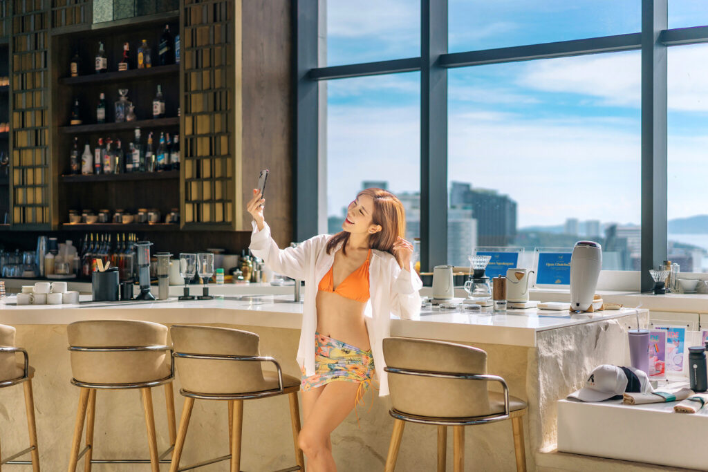 A woman in a bright outfit taking a selfie at a stylish coffee bar with a stunning view of Pattaya’s cityscape in the background.