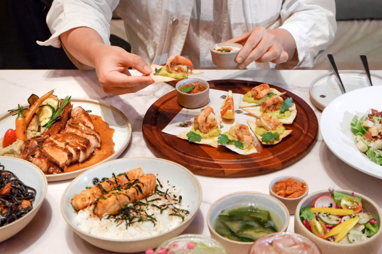 A variety of dishes served at a restaurant in Pattaya, featuring grilled chicken with sauce, shrimp tacos, rice with salmon, fresh salad, and soups, elegantly presented on a table.