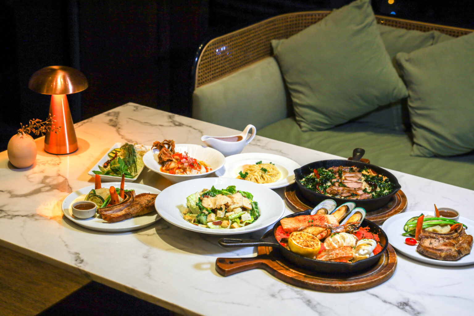 An elegant dinner spread featuring seafood paella, grilled meats, pasta, salad, and roasted vegetables on a marble table with warm ambient lighting.