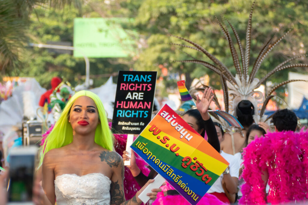 A participant in a Pride parade wearing a white dress and bright green hair, holding a colorful sign advocating for transgender rights. The background features other attendees in vibrant costumes and rainbow-themed signs.