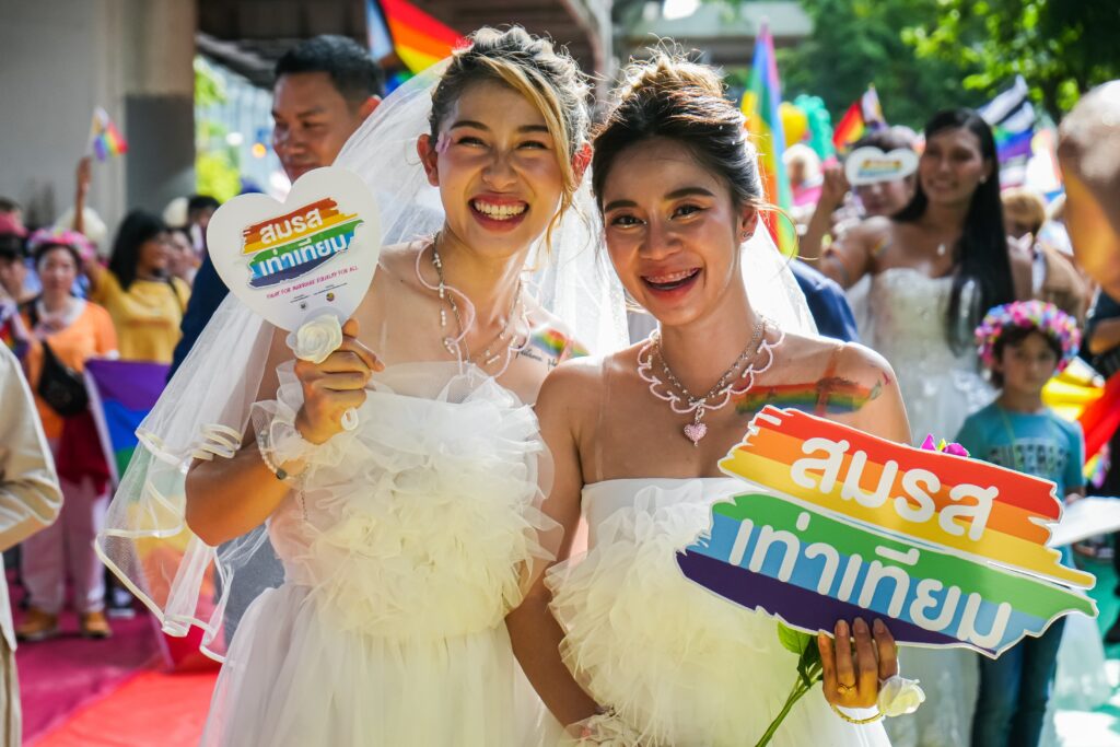 Two women dressed in white wedding-style outfits smile brightly, holding rainbow-themed signs advocating for marriage equality. They are surrounded by a colorful crowd during a Pride celebration.