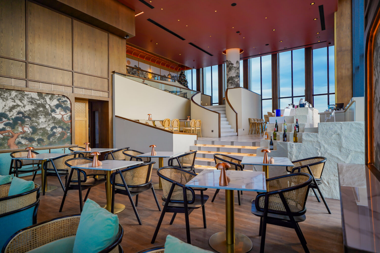 Modern restaurant interior showcasing a spacious two-level dining area with elegant marble tables, woven bistro chairs, and large windows providing ample natural light. The decor features warm wooden tones and a large mural, enhancing the contemporary yet cozy ambiance.