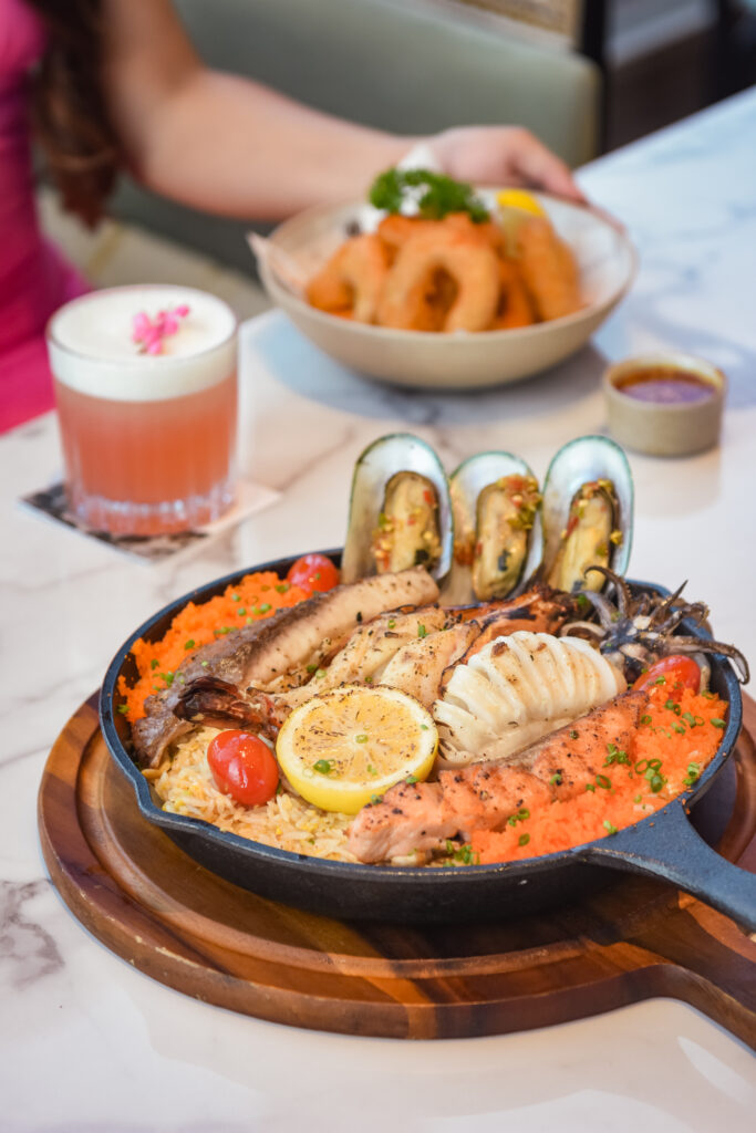 A delectable seafood platter featuring grilled salmon, squid, and mussels, served on a bed of seasoned rice with lemon slices and cherry tomatoes, accompanied by onion rings and refreshing beverages on a marble table.