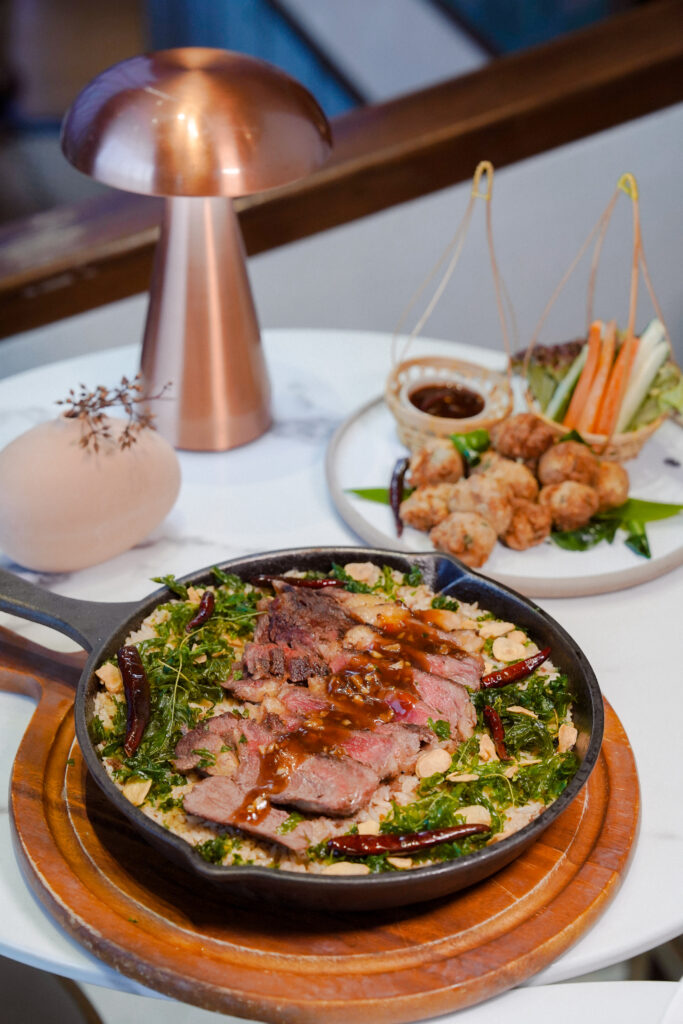 A gourmet meal featuring thinly sliced beef over a bed of crispy herbs in a skillet, accompanied by a side of fresh vegetables and Thai-style fried meatballs, presented elegantly on a marble table.