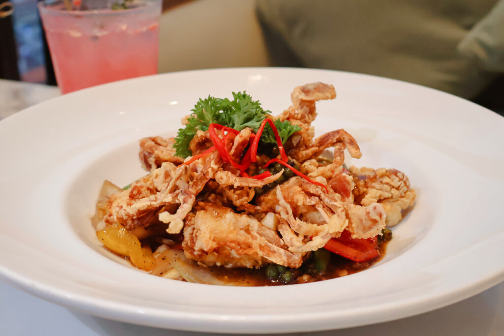 A beautifully plated dish featuring crispy soft-shell crab served over a medley of stir-fried vegetables, garnished with red chili strands and fresh parsley, accompanied by a pink cocktail in the background.