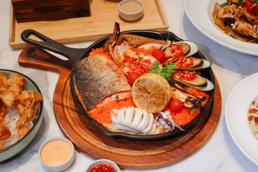 A vibrant seafood platter served at a restaurant, featuring a variety of seafood including grilled fish, shrimp, mussels, and squid, garnished with lemon and fresh herbs, presented on a sizzling hot skillet over a bed of bright orange rice.