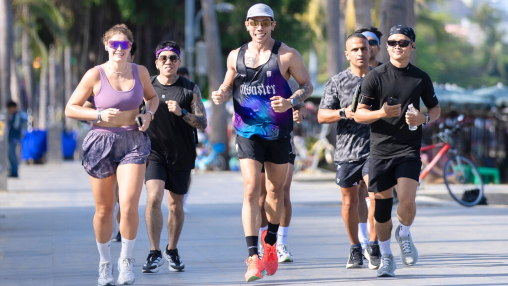A dynamic, sun-drenched photograph showing a diverse group of six people running together on a paved beachfront path in Pattaya. Integrated into the image are the stylized text: 'RUN TO YACHT PATTAYA PRIDE' and 'Kick off your wellness journey with a coastal city run led by Monster Run BKK along the vibrant beachfront.' This image illustrates the wellness and community aspect of the 'Run to Yacht' experience in Pattaya.