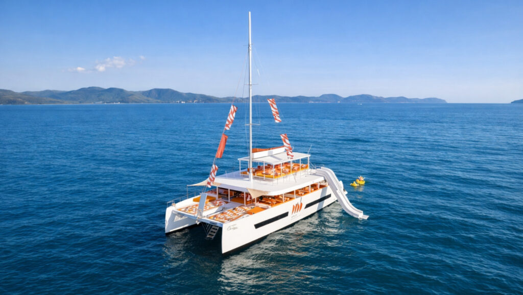 An aerial view of the luxury ONCE Yachting catamaran in Pattaya, with its distinctive orange and white theme, moored in the blue ocean with coastal hills. The boat features an inflated water slide and a small yellow floaty with people, showcasing the 'Run to Yacht in Pattaya' event experience.