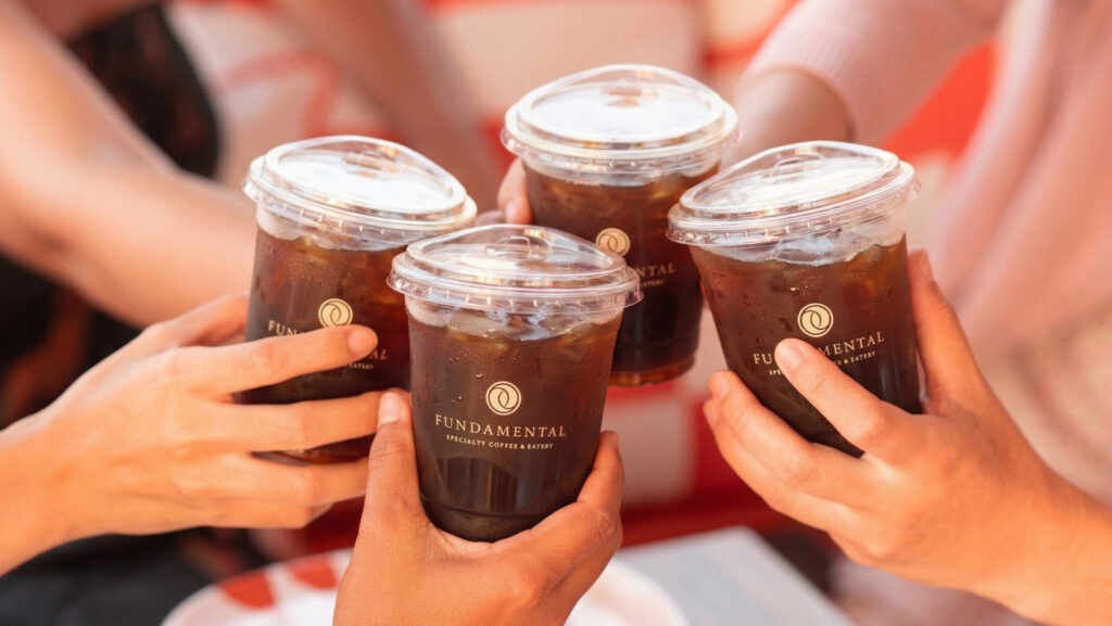 A close-up photograph of four diverse hands coming together in a toast, each holding a clear plastic takeaway cup filled with iced Americano coffee and ice. The cups are branded with the 'FUNDAMENTAL SPECIALTY COFFEE & EATERY' logo. The background shows blurred red and white striped outdoor seating under warm daylight.