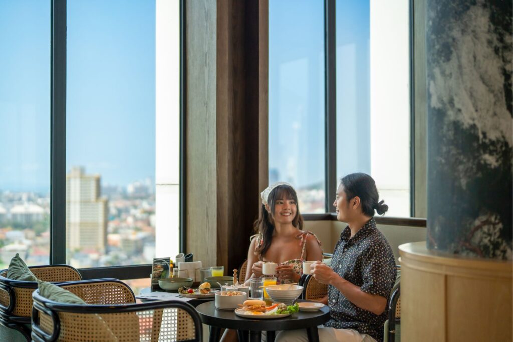 A couple enjoys breakfast and hot drinks beside floor-to-ceiling windows overlooking Pattaya at Arbour Hotel and Residence Pattaya.