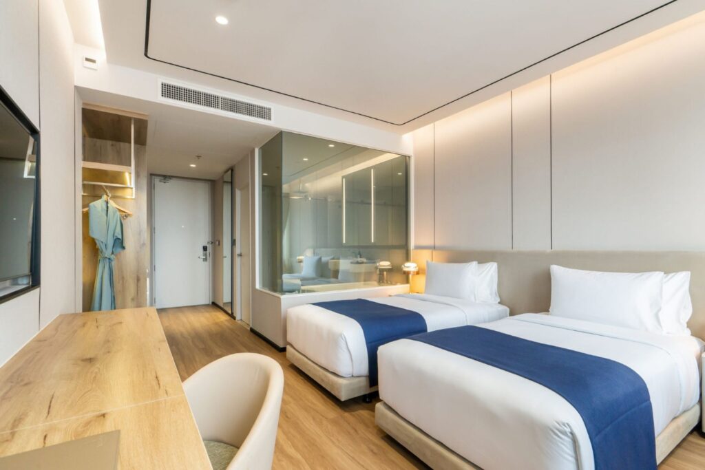 Bright twin-bed room at Arbour Hotel and Residence Pattaya with white bedding, blue runners, wooden desk, wardrobe, and glass bathroom.