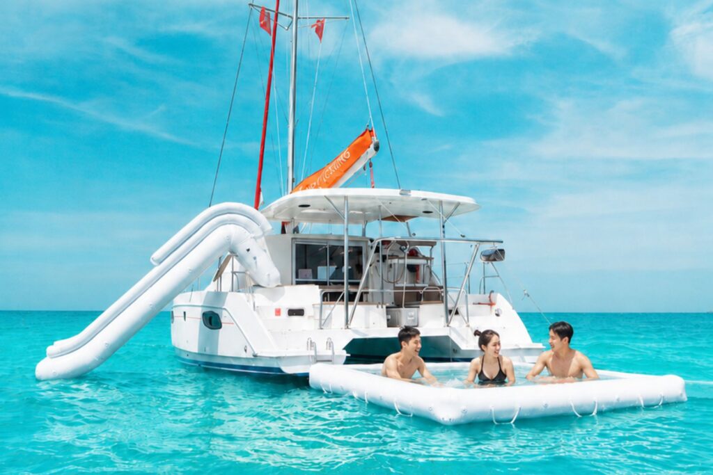 Three friends relax in a floating sea pool beside a white sailing catamaran equipped with a large inflatable water slide near Pattaya.