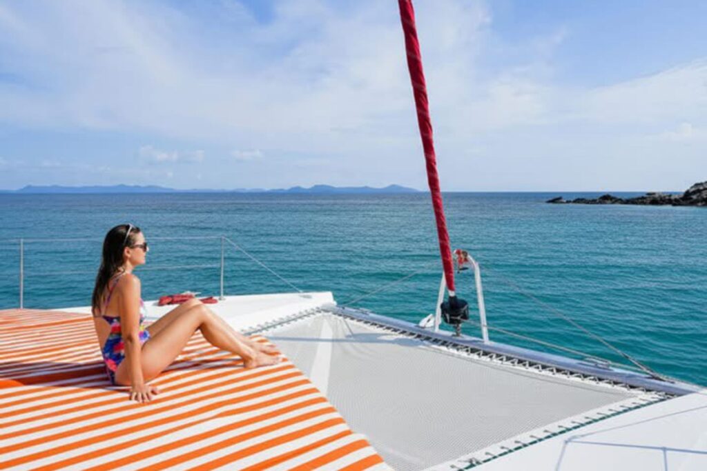 A woman in a colorful swimsuit relaxes on the front deck of a sailing catamaran while overlooking the calm blue sea near Pattaya.