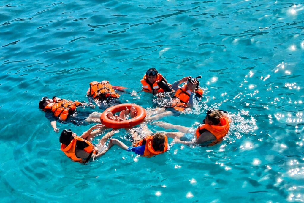 A group wearing orange life jackets and snorkeling masks floats together in clear turquoise water during a Pattaya yacht trip.