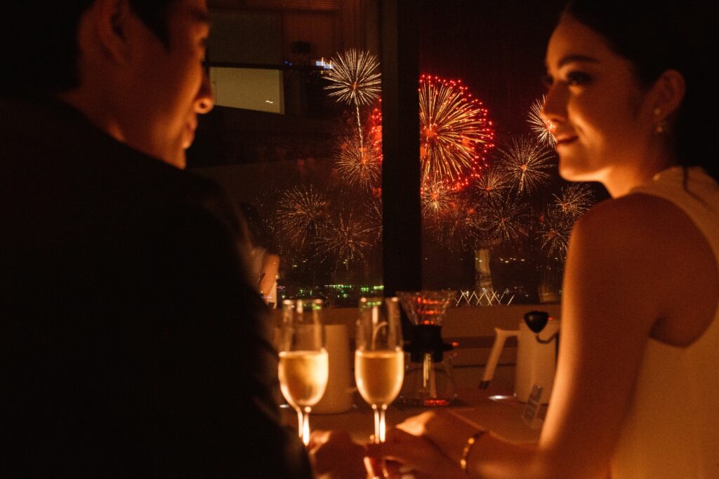 Couple enjoying dinner with champagne while colorful fireworks light up the night sky outside during the Pattaya Fireworks Festival.