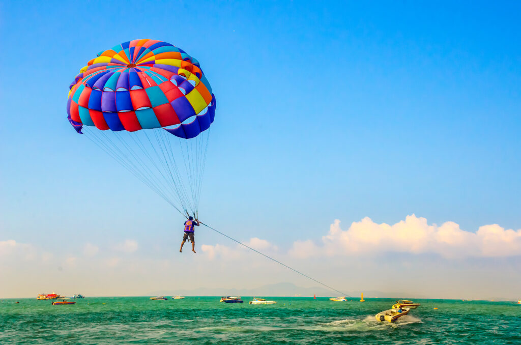A traveler wearing a life jacket enjoys parasailing above the sea in Pattaya beneath a colorful parachute, with boats visible on the water below.