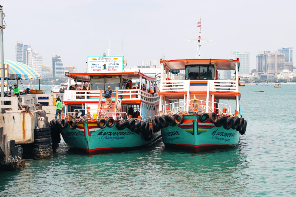 Two colorful passenger ferries dock at a Pattaya pier before traveling to Koh Larn, with the sea and Pattaya’s waterfront skyline behind them.