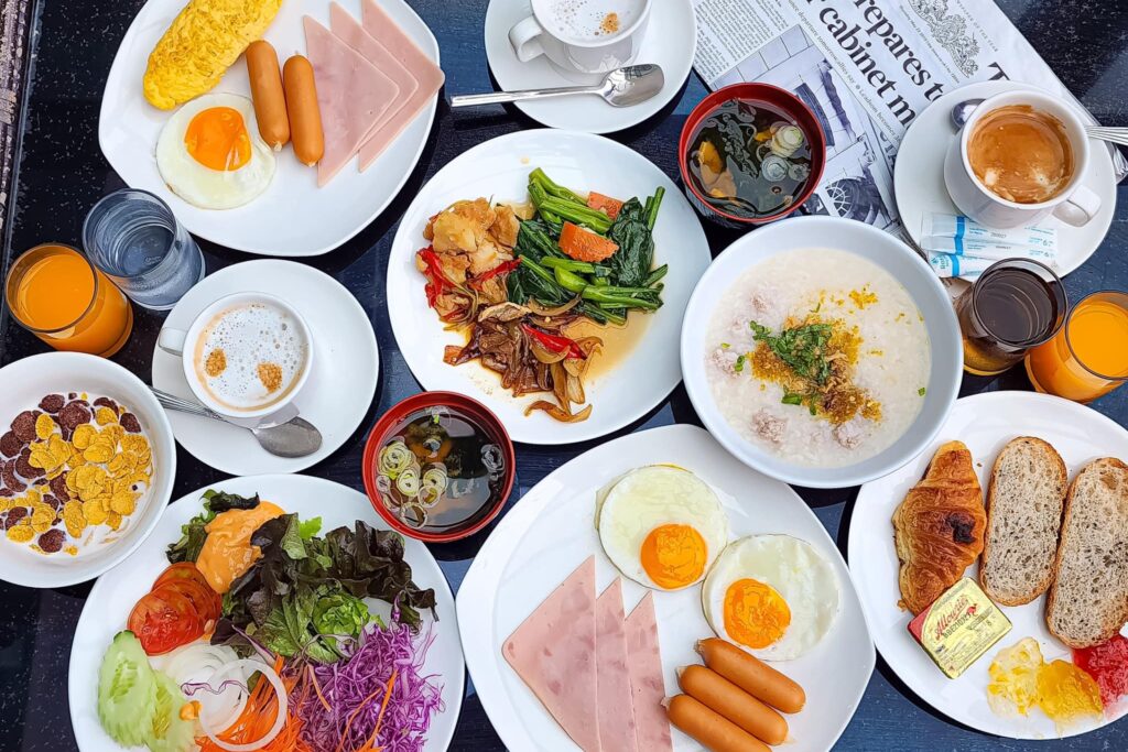 Breakfast spread at Arden Hotel and Residence Pattaya with eggs, sausages, ham, congee, vegetables, bread, cereal, coffee, and orange juice.