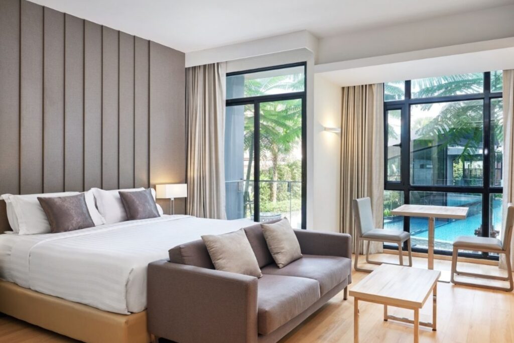 Spacious guest room at Arden Hotel and Residence Pattaya with a king bed, sofa, dining area, large windows, and views toward the swimming pool.