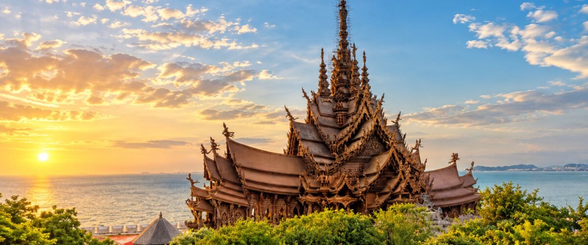 The Sanctuary of Truth in Pattaya rises above lush greenery beside the sea, illuminated by warm sunset light beneath a blue and golden sky.