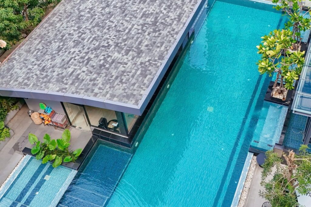 High-angle view of a large outdoor swimming pool at a Pattaya hotel, surrounded by tropical greenery, poolside seating, and modern buildings.