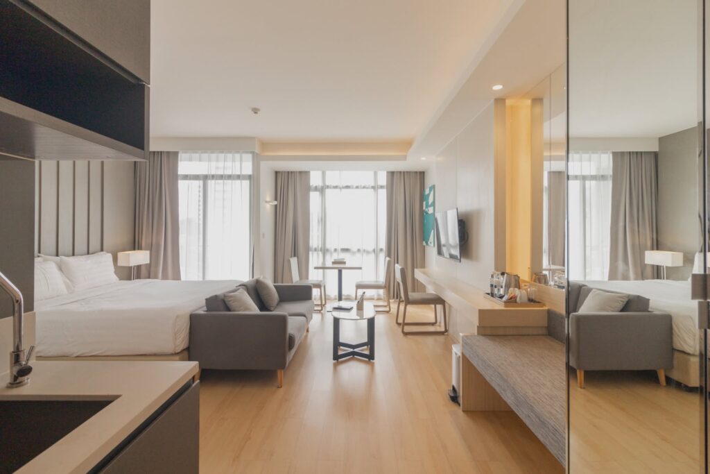 Spacious Pattaya hotel room with a double bed, sofa, coffee table, dining area, work desk, kitchenette, large mirror, and floor-to-ceiling windows.