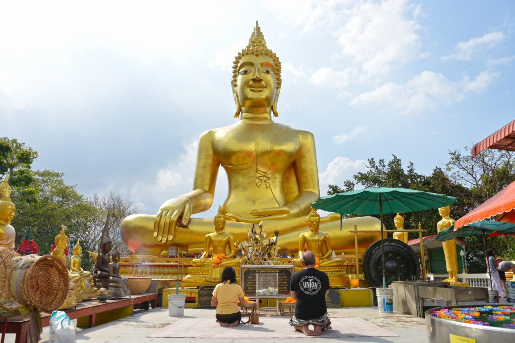 A golden Buddha statue at Wat Phra Yai (Big Buddha Temple) in Pattaya, Thailand, with two worshippers kneeling in front of the shrine under a bright sky.