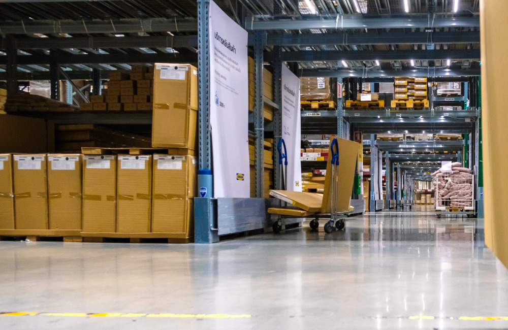 A spacious warehouse aisle inside IKEA Bangna with stacked boxes on shelves, a flat cart with packages, and industrial shelving.