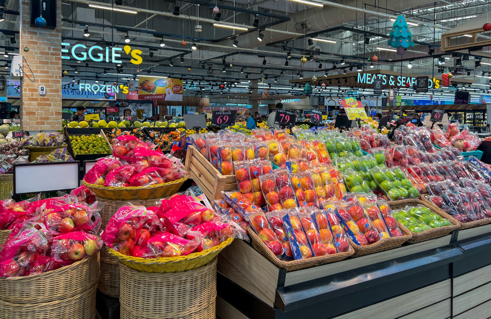 Fresh fruit display at Lotus's Sukhumvit 50 featuring neatly packed apples, grapes, and other produce, with signs for meats and seafood in the background.